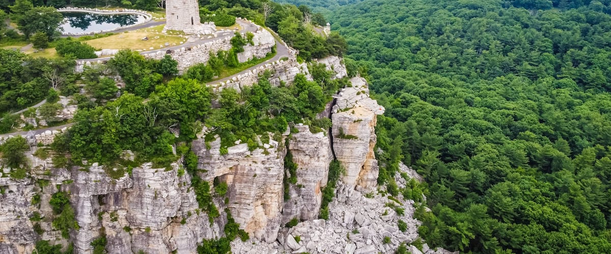 A stone tower on a cliff, surrounded by dense forest and a winding road.