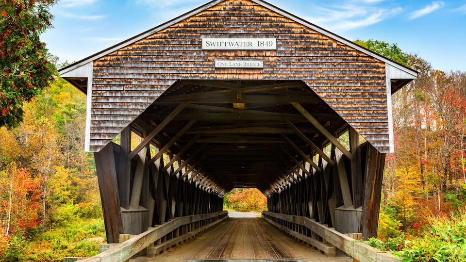 A covered wooden bridge labeled "Swiftwater 1849 One Lane Bridge" with a clear view of the surrounding autumn foliage.
