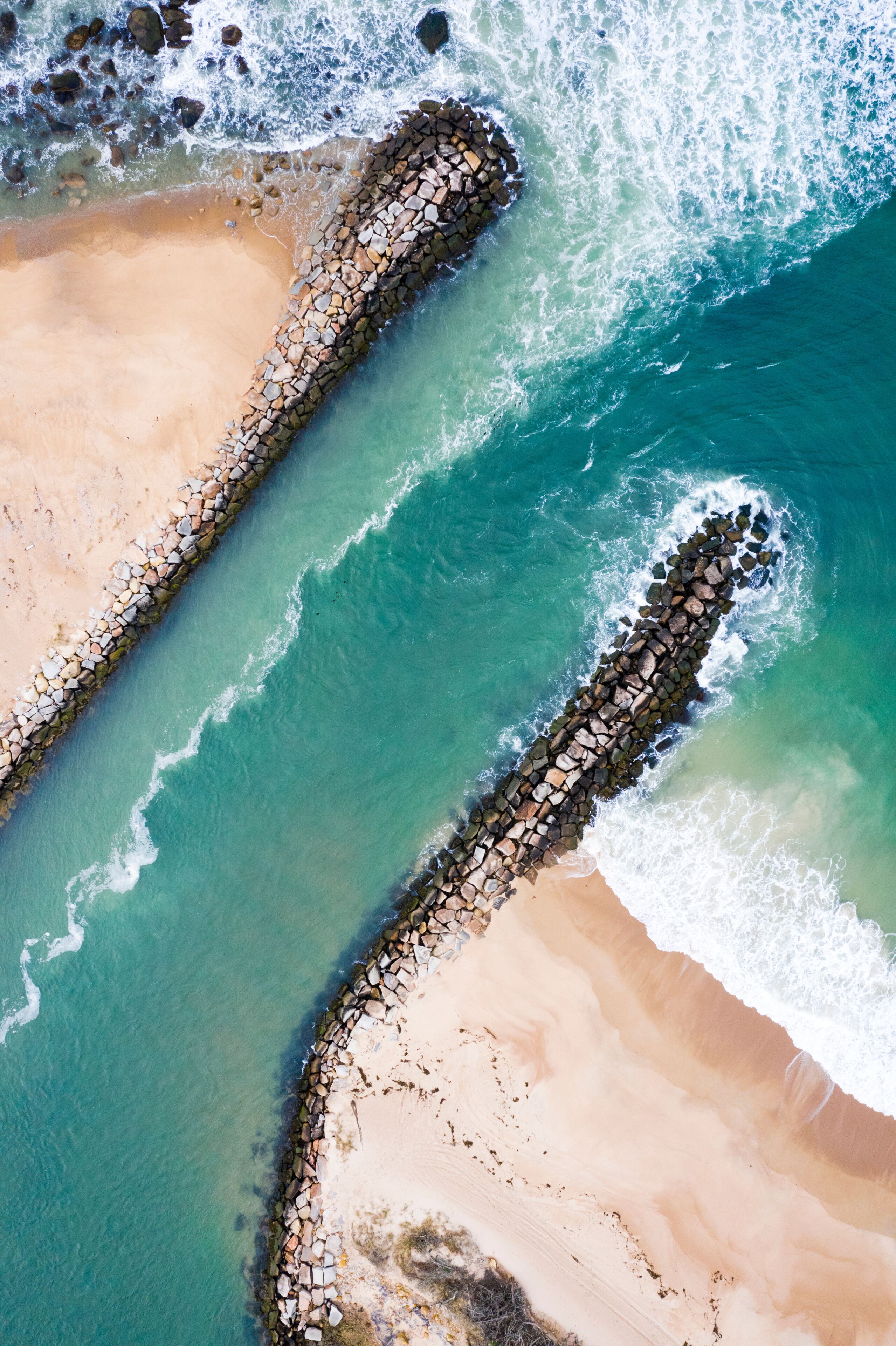 A coastal landscape with a stone jetty extending into the sea, clear turquoise waters, and sandy beach areas.