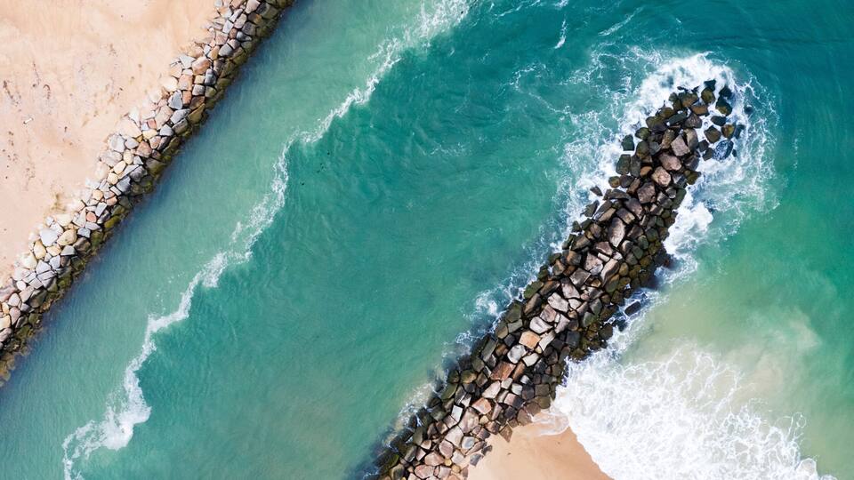 A coastal landscape with a stone jetty extending into the sea, clear turquoise waters, and sandy beach areas.