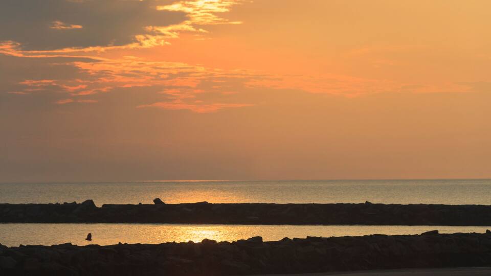 A beach at sunset with a silhouette of a person walking, the ocean, and a rocky shoreline.