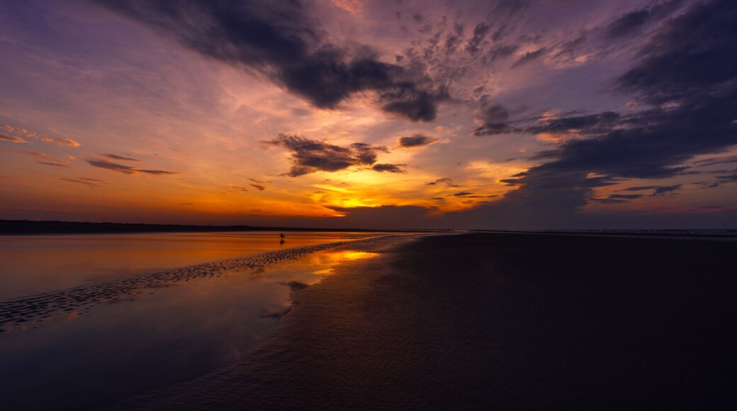 Un coucher de soleil sur une plage paisible, avec une personne qui se promène le long du rivage.