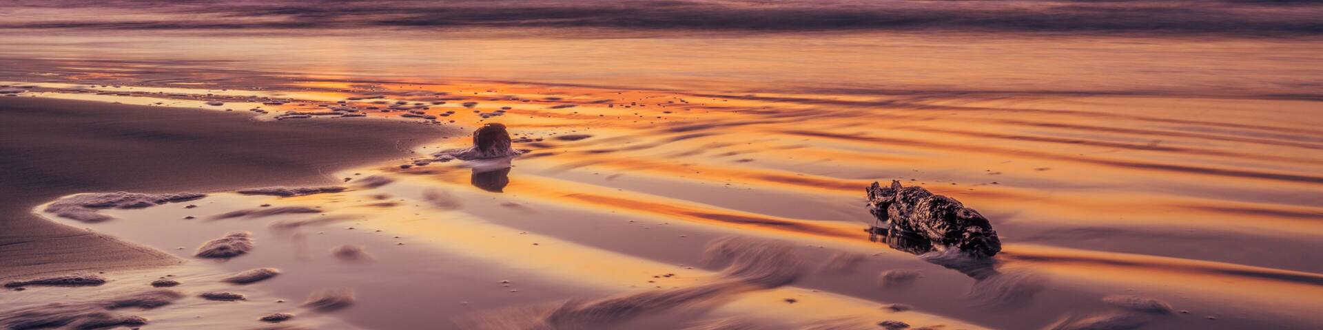 A beach at sunset with a calm sea, a rock formation, and footprints in the sand.