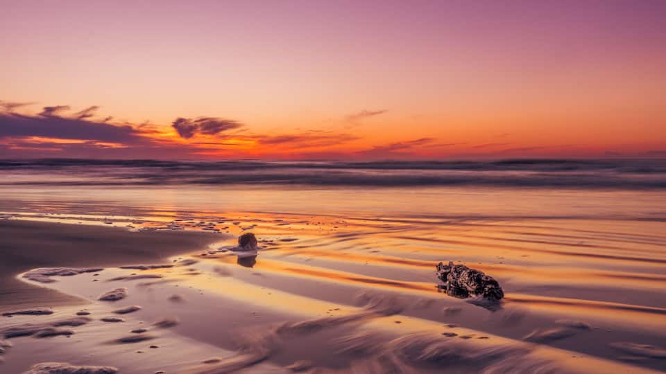 A beach at sunset with a calm sea, a rock formation, and footprints in the sand.