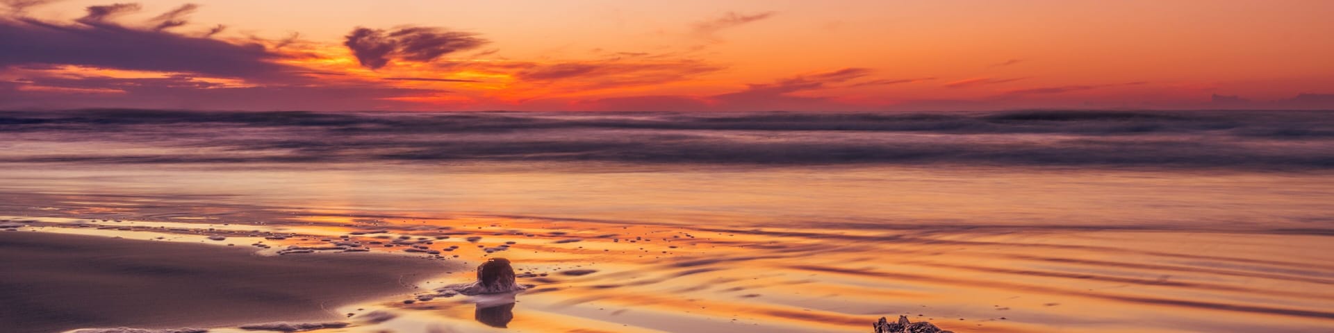 Une plage au coucher du soleil, avec une mer calme, une formation rocheuse et des empreintes de pas dans le sable.