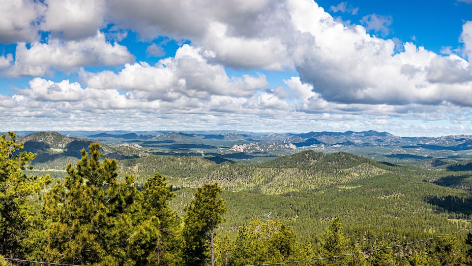 A mountainous landscape with dense forests and a clear blue sky with scattered clouds.
