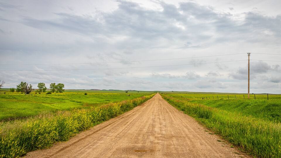 A dirt road in a rural area with green fields and a cloudy sky.