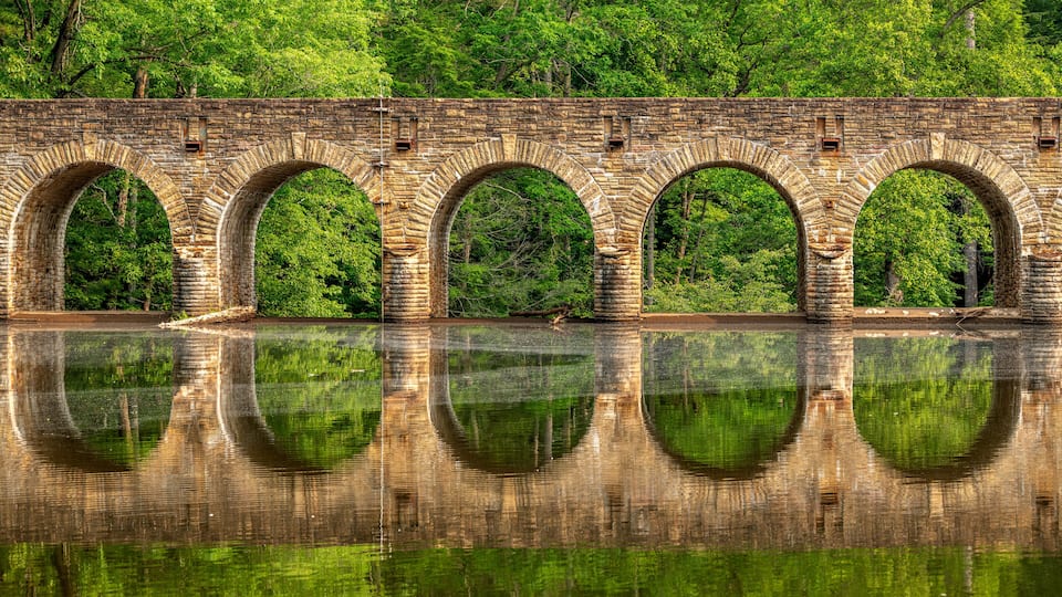 A stone bridge with multiple arches over a calm river, surrounded by lush green trees.
