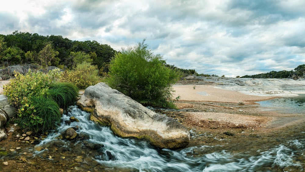 Ein Fluss fließt durch eine felsige Landschaft, mit einem Sandstrand und einem dichten Wald im Hintergrund.