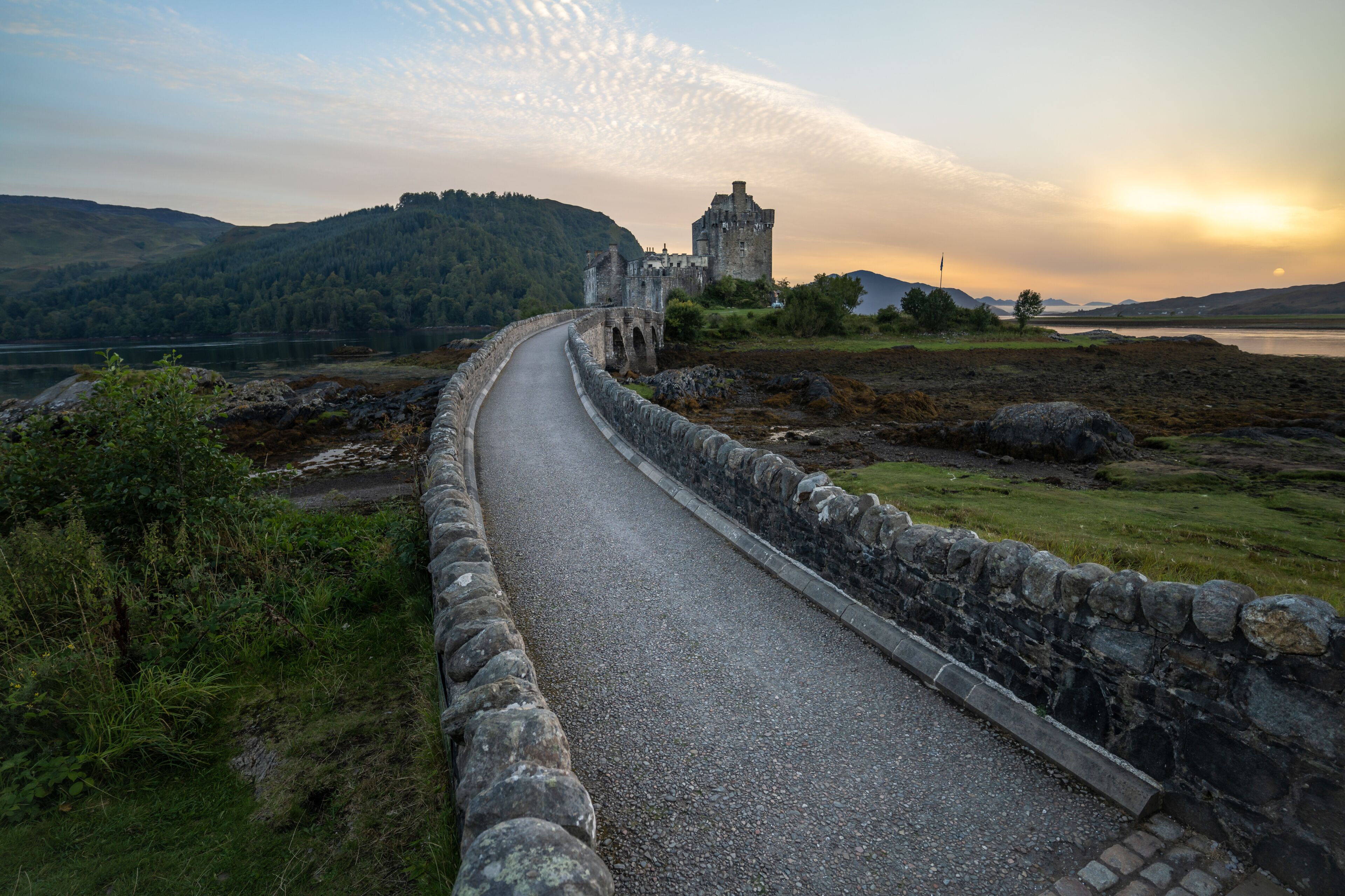 View of a stone bridge leading to a historic castle under a serene sky where the sun kisses the horizon, Eilean Donan Castle, Kyle of Lochalsh, Scotland, United Kingdom.