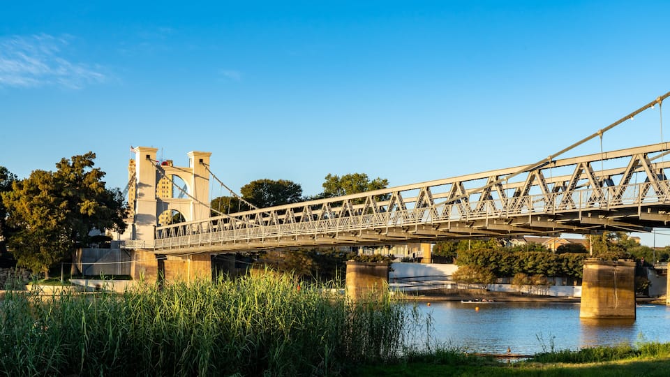 A steel bridge with towers, spanning over a river with greenery and trees in the background.