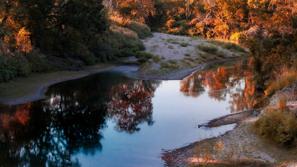 The Lampasas River in the Texas Hill Country, is framed by a sandy shoreline and lush autumn foliage. The vibrant orange, red, and yellow hues, along with their reflections in the calm water.