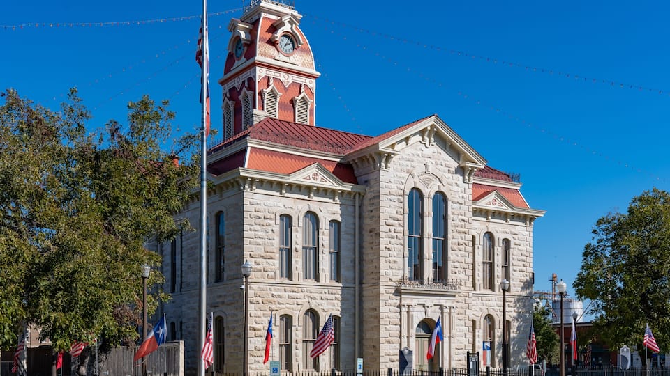 A historic stone building with a clock tower, surrounded by flags and trees.