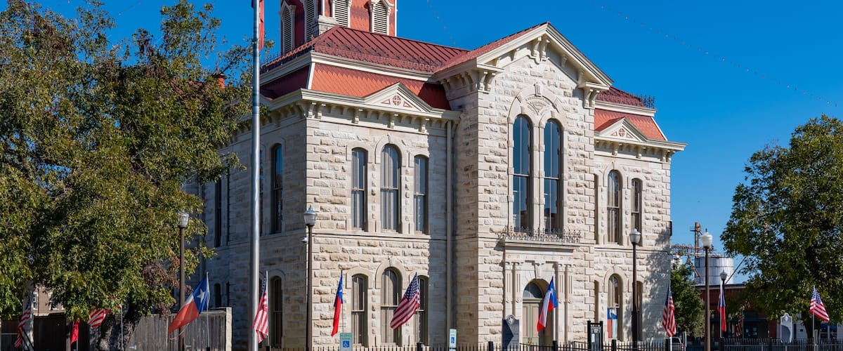 A historic stone building with a clock tower, surrounded by flags and trees.