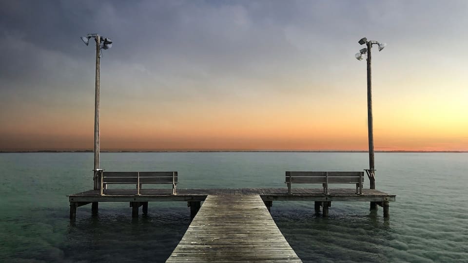 A wooden pier extending into calm water with two benches at each end, under a clear sky.