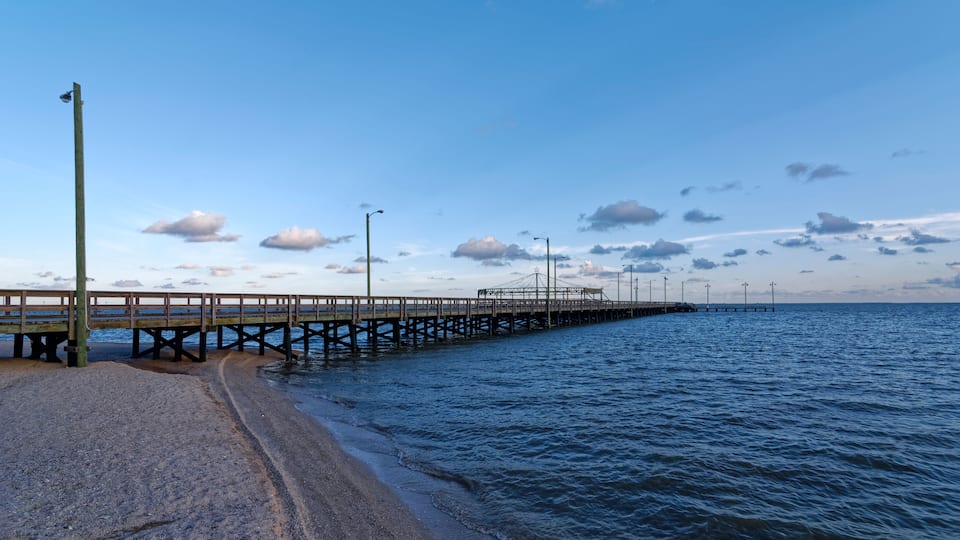 A beach with a wooden pier extending into the ocean, under a partly cloudy sky.