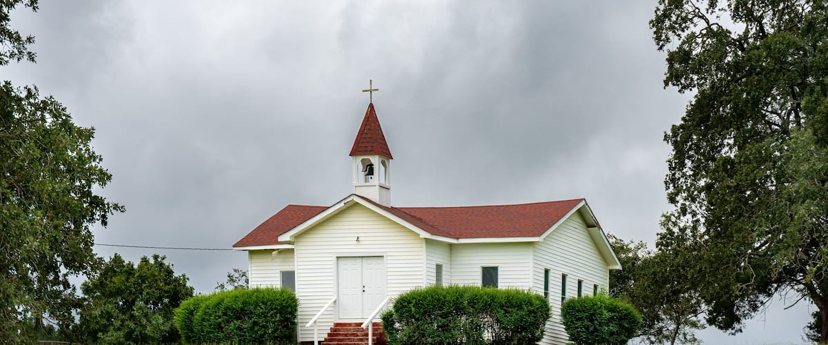 A white church with a red roof and a cross on top, surrounded by green grass and trees.
