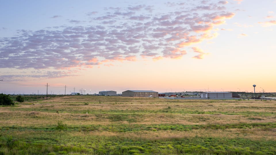 A grassy field with industrial buildings and power lines in the background.