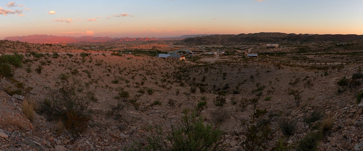 A desert landscape with sparse vegetation, scattered buildings, and distant mountains under a clear sky.