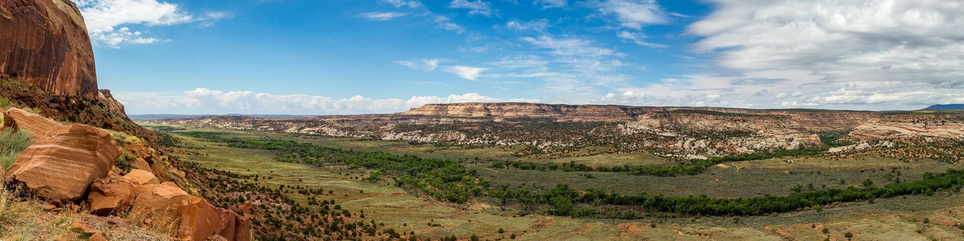 A vast landscape with a prominent red rock formation on the left, a green valley, and layered hills in the distance under a partly cloudy sky.