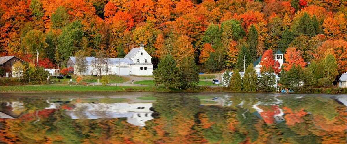 A lake with autumn foliage reflecting on the water, surrounded by white buildings and a forested hillside.