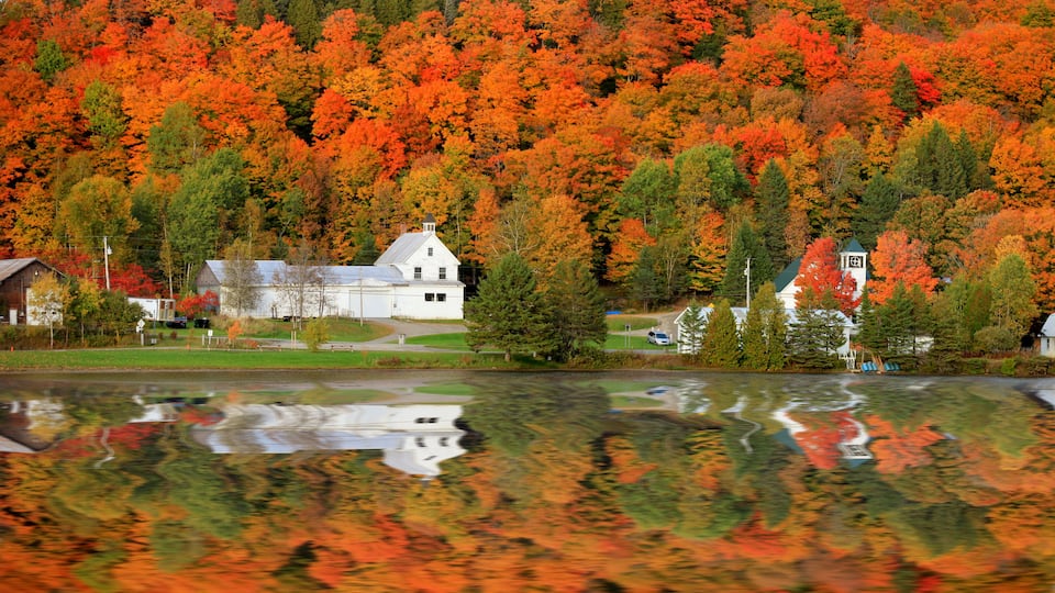 A lake with autumn foliage reflecting on the water, surrounded by white buildings and a forested hillside.