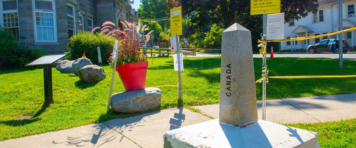 A stone monument with the inscription "CANADA" surrounded by greenery, a red planter, and a building in the background.