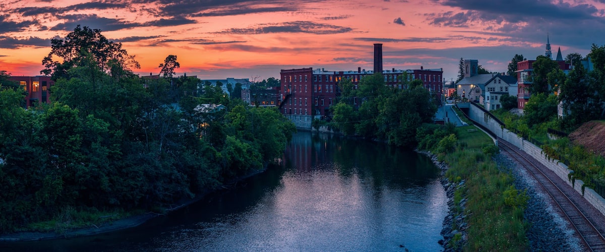 A river with a bridge, a train track, and buildings with a church steeple in the background.