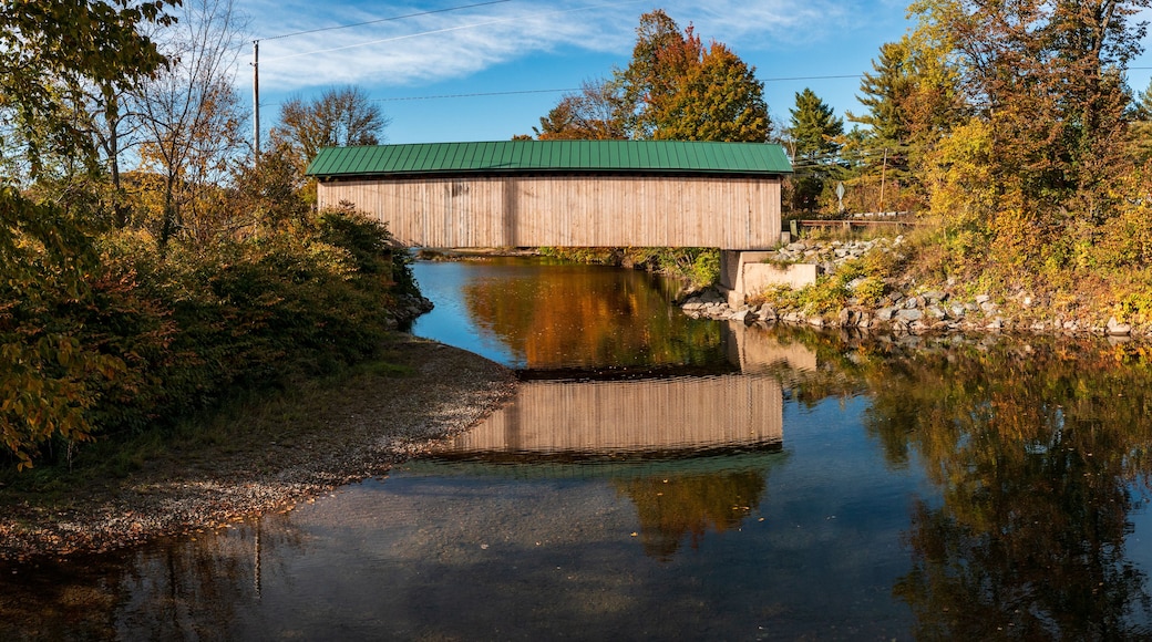 A wooden covered bridge over a calm river, surrounded by autumn foliage.