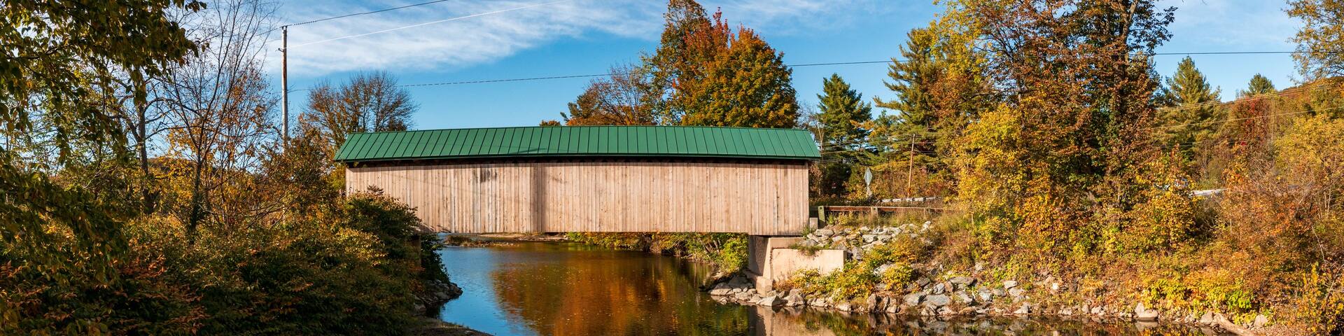 A wooden covered bridge over a calm river, surrounded by autumn foliage.