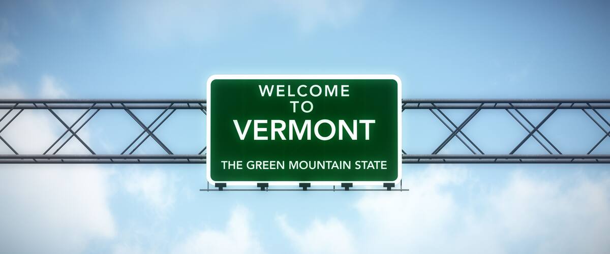 A road sign with 'WELCOME TO VERMONT' and 'THE GREEN MOUNTAIN STATE' against a sky with clouds.