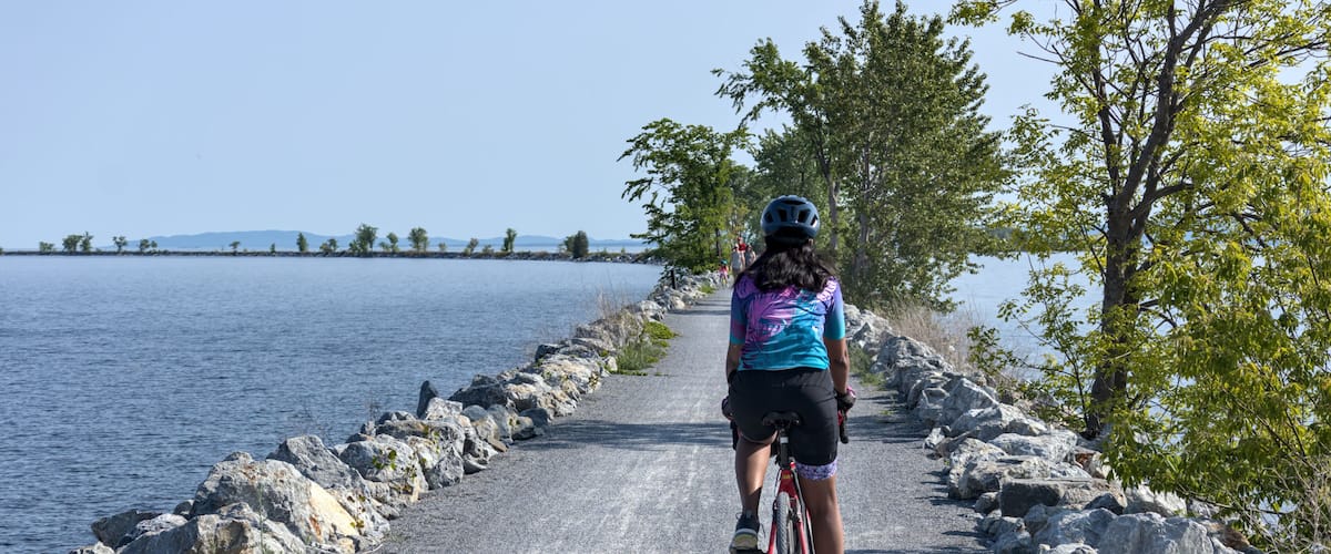 A cyclist on a gravel path by a lake with rocky edges and trees.