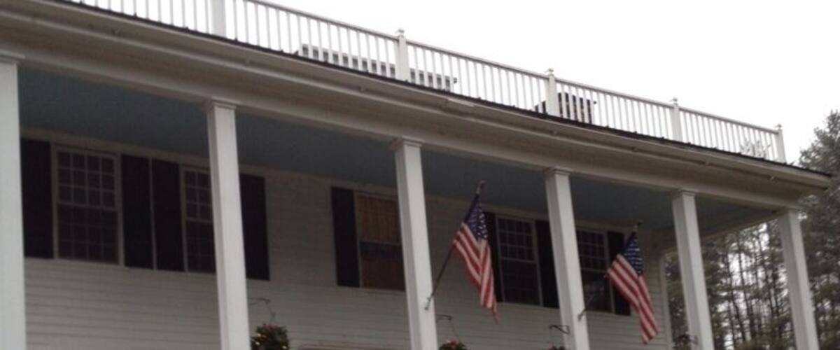 A two-story white house with a porch, American flags, and a stone wall.