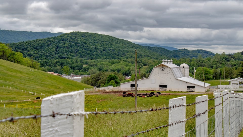 A white barn with a clock, surrounded by green fields and mountains in the background.