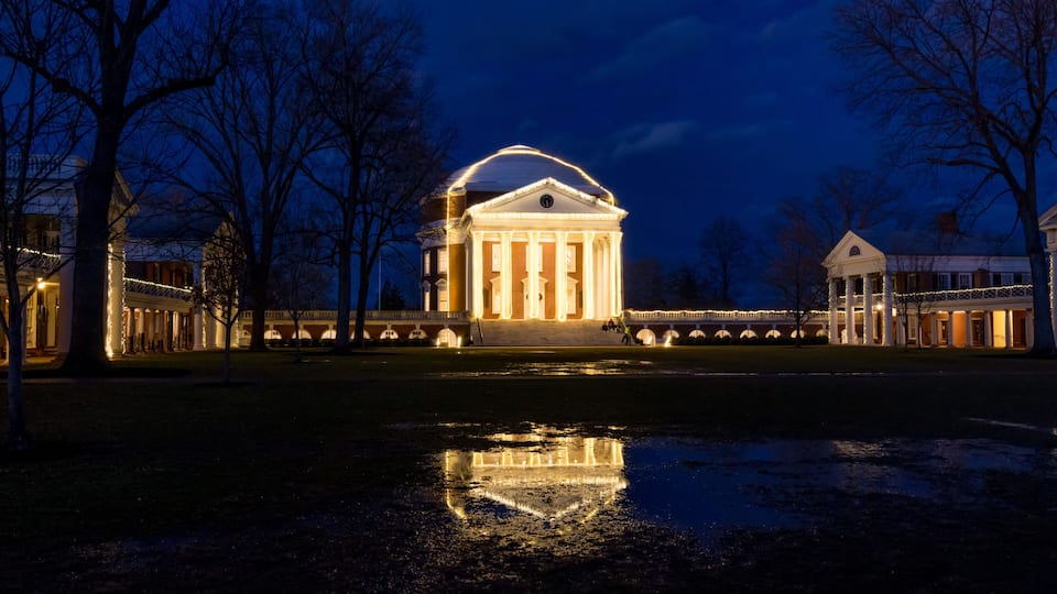 A classical building with a dome, surrounded by trees and a body of water reflecting its lights.