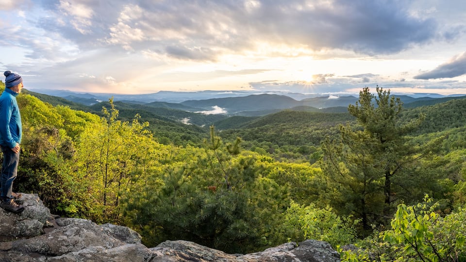 A person standing on a rocky outcrop overlooking a mountainous landscape with dense forests and a valley below.