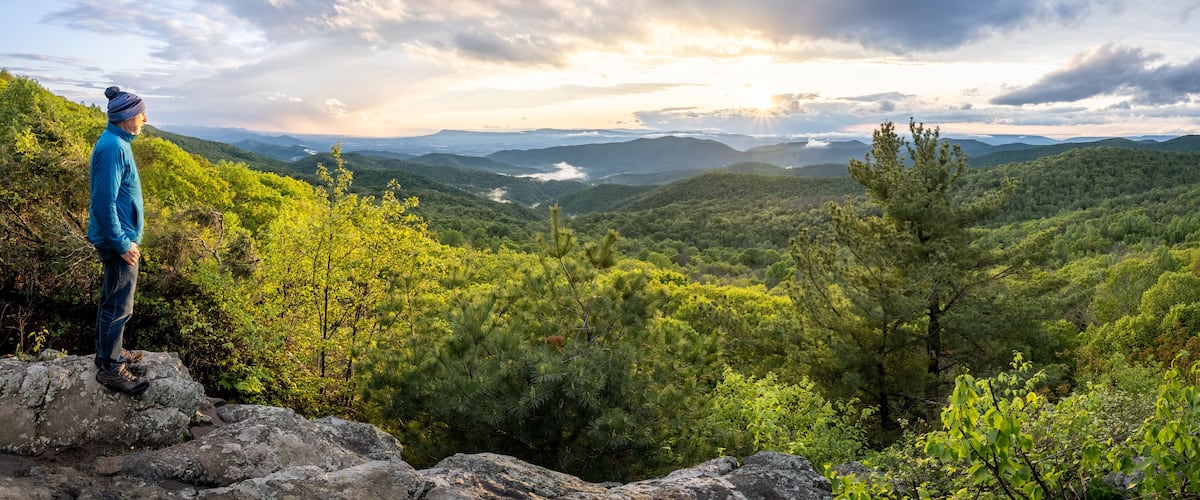 A person standing on a rocky outcrop overlooking a mountainous landscape with dense forests and a valley below.