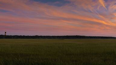 A sunset over a grassy field with a lighthouse in the distance.