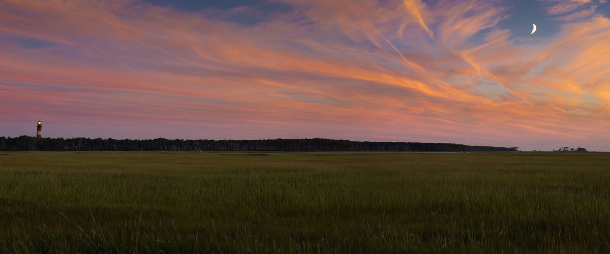 A sunset over a grassy field with a lighthouse in the distance.