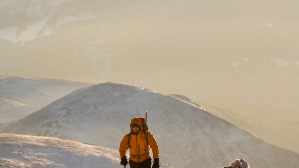 A hiker in an orange jacket on a snowy mountain with a clear sky.