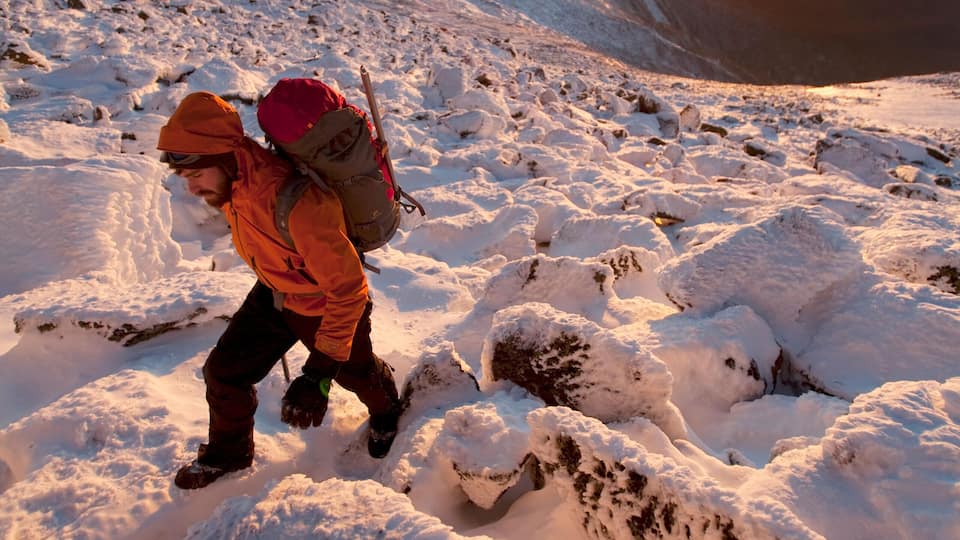 A hiker in an orange jacket and hat with a red backpack on a snowy mountain path during sunset.