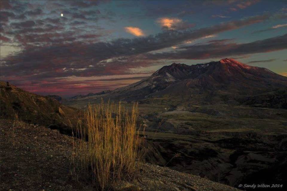 Una montaña con un resplandor rojizo en la cima, rodeada por un valle de vegetación escasa.