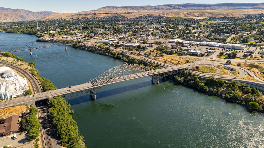 Une rivière traversée par un pont, une ville et des montagnes en arrière-plan.
