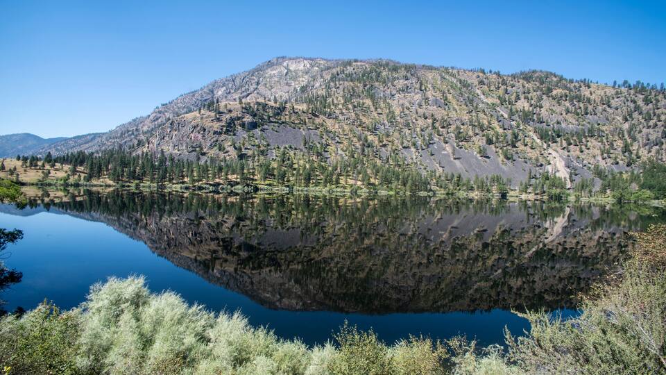 A mountain with a lake reflecting its image, surrounded by trees and shrubs.