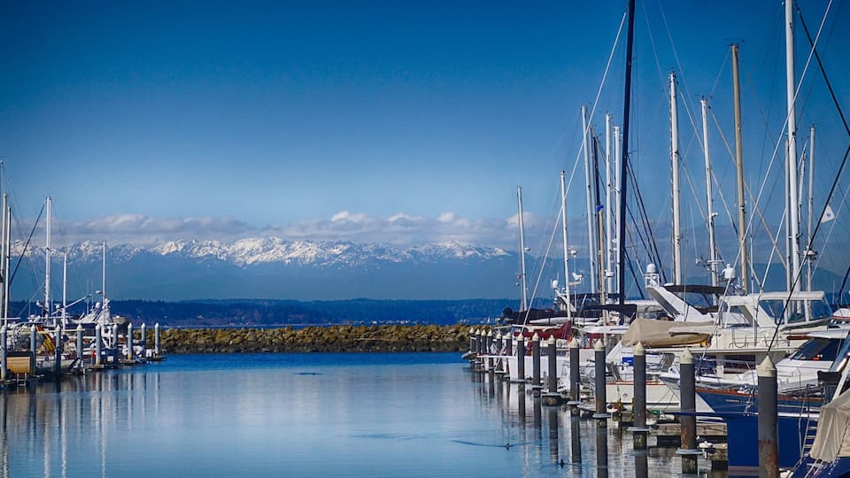 A marina with boats docked, a calm water surface, and snow-capped mountains in the background.