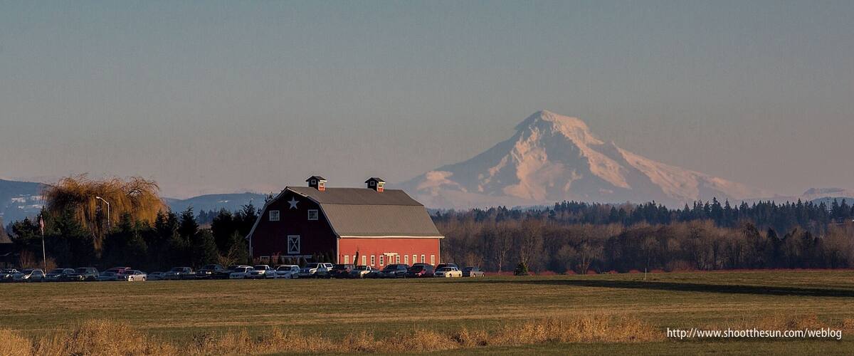 A red barn with a star on the roof, a parking lot with cars, and a snow-capped mountain in the background.