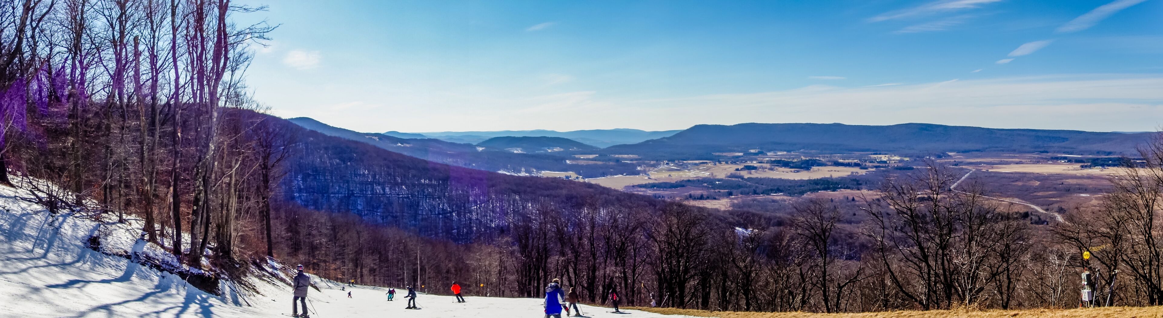 A snowy ski slope with skiers, a mountain range, and a valley.