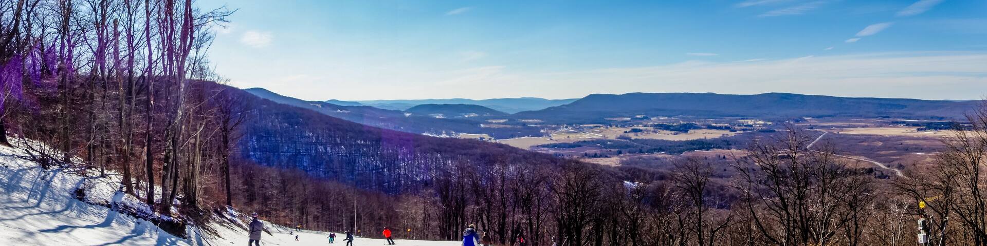 A snowy ski slope with skiers, a mountain range, and a valley.