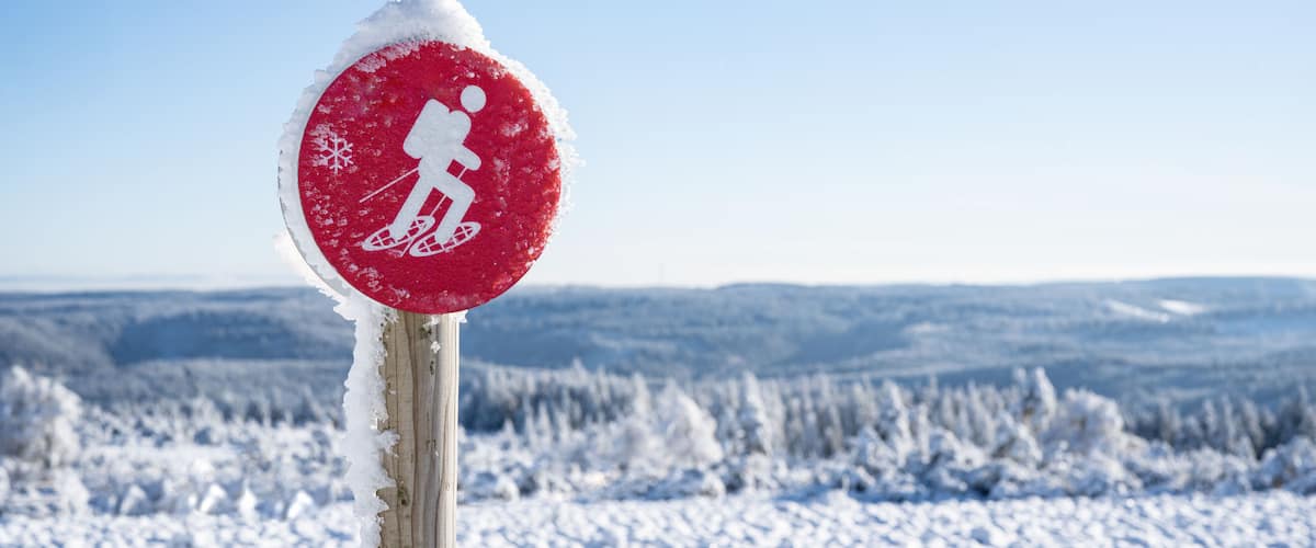 A red sign with a skier symbol, surrounded by snow, with a snowy landscape in the background.