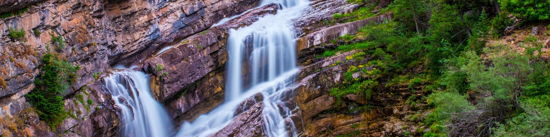 A multi-tiered waterfall cascading down a rocky cliff surrounded by lush greenery.